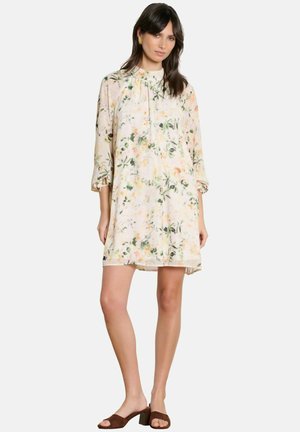 Naine kandes helelillelist põlvpikkust kleiti, millel on kolmveerandvarrukad, koos pruunide plokktaldsete avatud varvastega sandaalidega, seistes valge taustaga ees.