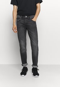 Jeans de denim cinza escuro com um corte justo, apresentando cinco bolsos, desbotamento leve e punhos arrolados. Usados com sapatilhas de atletismo pretas.