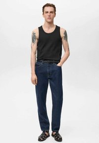 Zwarte tanktop, blauwe spijkerbroeken en zwarte sandalen. De spijkerbroek is losvallend met een klassieke snit. Tattoos zichtbaar op de armen.