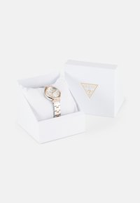 Montre en argent et or rose présentée dans une boîte blanche, avec un cadran rond, un bracelet en métal et un logo sur le cadran, reposant sur un coussin blanc.