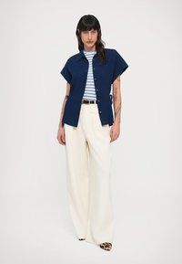 Μπλε πουκάμισο με κοντά μανίκια, φορεμένο πάνω από μπλε και λευκή ριγέ μπλούζα, συνδυασμένο με λευκό παντελόνι με φαρδιά μπατζάκια και παπούτσια με λεοπάρ μοτίβο.