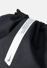 Fekete húzózsinóros táska textúrált anyagból, fehér címkével, rajta szöveggel. Fekete Nike logóval rendelkezik.