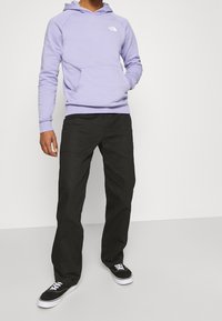 Persoon in een lichtpaarse hoodie, zwarte broek en zwarte sneakers met witte veters tegen een effen witte achtergrond.