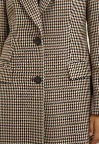 Hnedý, čierny a biely kabát s károvaným vzorom houndstooth, dvoma čiernymi gombíkmi a klopovými vreckami, nosený osobou s viditeľnou pravou rukou.