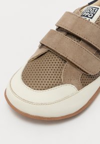 Zapatillas marrones y crema con parte superior de ante, textura perforada y dos correas de Velcro. Presenta una suela de goma y una etiqueta con marca.