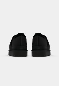 Paire de chaussures basses noires montrées de dos avec semelles plates et logo visible de la marque UGG sur la zone du talon.