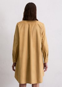 Persoon met schouderlang donker haar, die een losse, lange mouwen, knielange beige jurk draagt, tegen een eenvoudige witte muur staat.