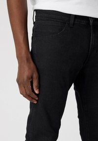 Vaqueros de denim negro con un corte ajustado, que cuentan con un diseño de cinco bolsillos, costuras visibles y una textura suave. La mano se apoya en la zona de la cadera.