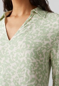 Chemise vert clair avec un motif abstrait blanc, dotée d'un col et d'un décolleté en V. Fabriquée en tissu texturé, idéale pour une tenue décontractée.