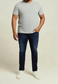Man som bär en ljusgrå t-shirt med kort ärm, mörkblå jeans och vita sneakers, står med en hand i fickan mot en enfärgad bakgrund.