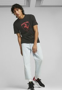 Czarna koszulka z logo Ferrari w kolorze czerwonym, zestawiona z jasnoniebieskimi dżinsami i czarnymi trampkami. Materiał wygląda na gładki i ma dopasowany krój.