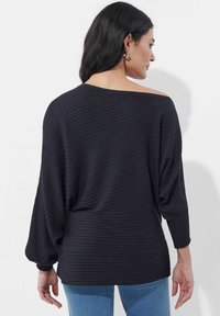 Schwarzer, gerippter Pullover mit schulterfreiem Design, langen Ärmeln und lockerem Schnitt. Endet auf Hüfthöhe und wird mit blauen Jeans getragen.