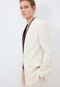 Kremfärgad blazer med randig mönster och en strukturerad design, med en enkel bröstficka, spetsigt slag och en avslappnad passform, bärs över en mörk topp.