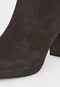 Gros plan d'une botte chelsea en daim marron foncé avec un bout arrondi et un talon bloc sur un fond clair.