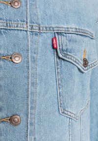 Světle modrá džínsová bunda se stříbrnými knoflíky, oranžovým prošíváním, výraznou přední kapsou a červenou nášivkou Levi's na bočním švu.