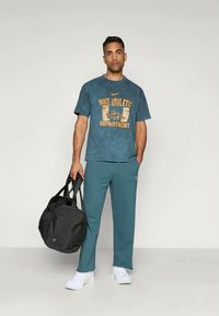 T-shirt desportivo da Nike em azul com texto amarelo, combinado com calças desportivas em verde-água. O modelo segura uma mala de duffle preta, vestindo sapatos brancos.