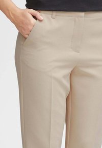 Pantaloni beige con frontale piatto, dettagli sulle tasche laterali e una texture liscia. Il tessuto sembra leggero e su misura.