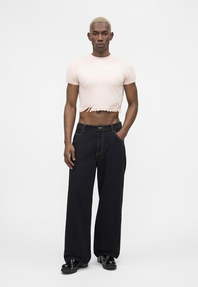 Άνδρας μοντέλο με κοντά ξανθά μαλλιά, φοράει ένα ανοιχτό ροζ κοντό t-shirt, μαύρο φαρδύ παντελόνι και μαύρα παπούτσια με πλατφόρμα, στέκεται μπροστά σε λευκό φόντο.