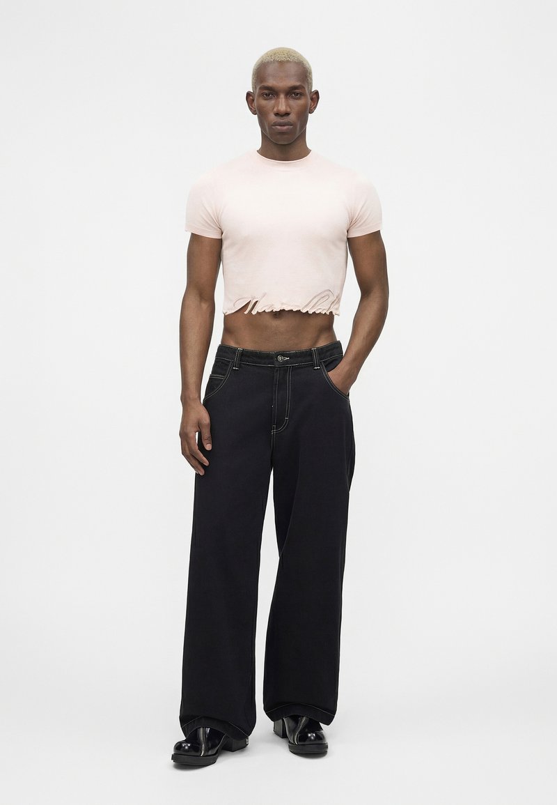 Mannequin masculin aux cheveux blonds courts portant un t-shirt court rose pâle, un pantalon large noir et des chaussures plateformes noires, se tenant devant un fond blanc.
