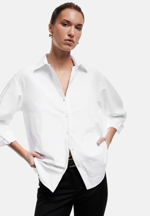 Camicia bianca con colletto strutturato e spalle scese, caratterizzata da una vestibilità ampia e una fila di bottoni visibile. Abbinata a pantaloni neri.