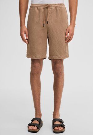 Mężczyzna w beżowych szortach ze ściągaczem i czarnych sandałach z klamrą, stojący na jednolitym jasnym tle.