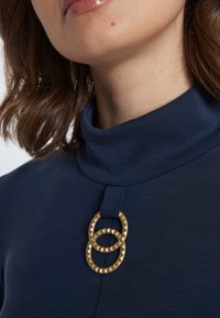 Dunkelblauer Rollkragenpullover mit einem strukturierten, goldenen ovalen Akzentdetail am Halsausschnitt. Glatte Stoffqualität, schlichtes Design und enganliegende Passform.