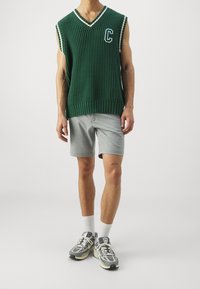 Zelený pletený svetr bez rukávů s výstřihem do V a bílými akcenty, doplněný o šedé šortky a multi-barevné běžecké boty.