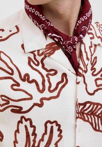 Camisa de manga corta en color blanco roto con patrones abstractos de hojas marrones. La tela tiene un acabado texturizado y ligeramente arrugado. Pañuelo de paisley marrón alrededor del cuello.