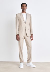 Beige Leinenanzug mit tailliertem Sakko und passender Hose, kombiniert mit einem weißen T-Shirt und weißen Sneakern. Glatte Textur, klassisches Design.