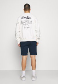 Valkoinen puuvillainen collegepaita, jossa musta graafinen painatus, jossa on leijona ja teksti, yhdistetty tummansinisiin shortseihin. Mallilla valkoiset tennarit.