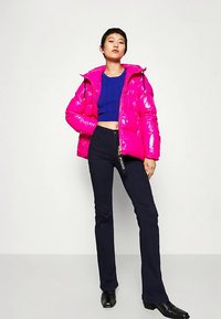 Žiarivá ružová lesklá nadýchaná bunda s kapucňou, na zips vpredu a s elasticnými manžetami, kombinovaná s modrým krátkym topom a čiernymi rozšírenými nohavicami.