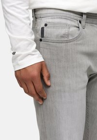 Jean en denim gris de coupe classique, avec des poches avant, une quincaillerie noire, et une chemise à manches longues blanche avec un logo sur la manche.