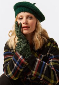 Guanti con stampa a leopardo verde, berretto berretto verde in lana, cappotto scozzese nero con strisce rosse, blu e verdi; i modelli testurizzati aumentano l'appeal visivo.