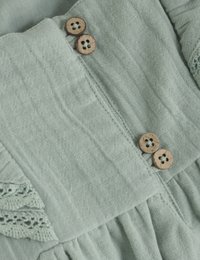 Tissu vert clair, texturé avec des plis et des accents en dentelle. Trois boutons en bois embellissent le devant, ajoutant une touche naturelle.