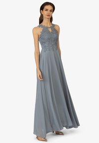 Langes, fließendes blaues Abendkleid mit Spitzenoberteil und Schlüsselloch-Detail. Verfügt über ein ärmelloses Design und einen weichen, strukturierten Stoff. Sichtbare akzentuierte Schuhe.