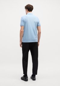 Gai zils polo krekls ar īsām piedurknēm, apvienots ar melniem sašaurinātiem biksēm un melnām kurpēm, redzams no aizmugures pret vienkāršu fona.