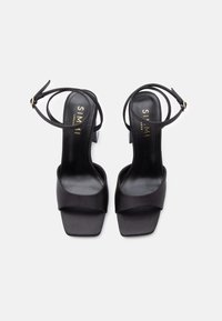 Melnas augstpapēžu sandales ar satīna apdari, platām priekšējām siksnām, potītes siksnu un kvadrātveida purngalu dizainu. Zelta krāsas zīmols zolēs.