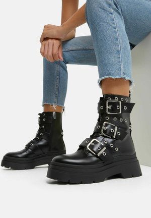 Persoon draagt lichtblauwe cropped spijkerbroek en zwarte leren combatlaarzen met drie zilveren gespen en dikke zolen, zittend met gevouwen handen.