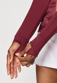 Top lungo bordeaux con fori per il pollice, realizzato in tessuto elastico; presenta un design aderente e dettagli lievi nelle cuciture, abbinato a una gonna bianca.