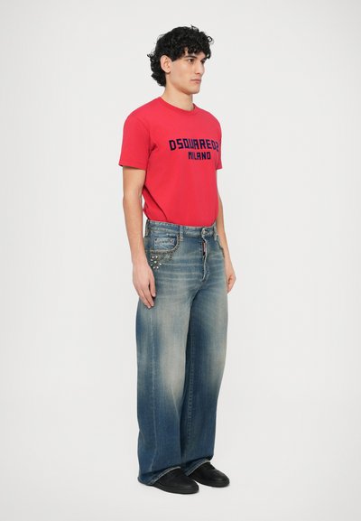 Červené tričko s námorníckym modrým nápisom, doplnené o široké vyblednuté modré džínsy s dekoračným reťazovým detailom na vrecku. Čierne topánky dopĺňajú vzhľad.