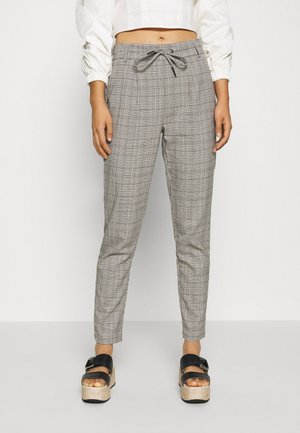 Vrouw die grijze geruite taps toelopende broek draagt met een taillekoord en zwarte plateauzandalen, staand tegen een effen witte achtergrond.