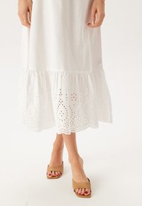 Vit bomullsklänning med en flikig fåll som har hålsöm och en scalloped kant. Matchad med bruna öppna sandaler.