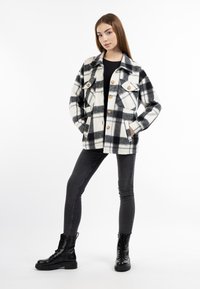 Giacca a quadri bianca e nera con due tasche sul petto, bottoni in legno e texture del tessuto. Indossata sopra una maglietta nera e abbinata a jeans skinny neri e stivali.