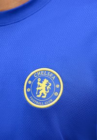 Μπλε αθλητική φανέλα με κίτρινο έμβλημα της Chelsea Football Club που απεικονίζει ένα λιοντάρι που κρατά ένα ραβδί, δύο ποδοσφαιρικές μπάλες και δύο λουλούδια.