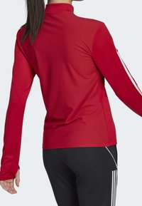 Frau in einem roten langärmeligen Sportoberteil mit strukturiertem Muster und schwarzen Hosen mit weißen Streifen, von hinten betrachtet.