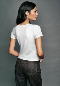 White short-sleeve fitted t-shirt with a round neckline, worn with dark brown textured pants featuring visible stitching and back pockets.