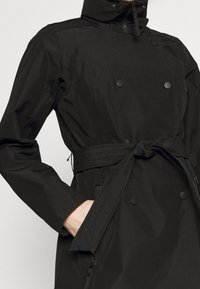 Trench coat impermeabile nero con colletto alto, design a doppia abbottonatura, vita con cintura e tasche laterali con zip per una maggiore funzionalità.