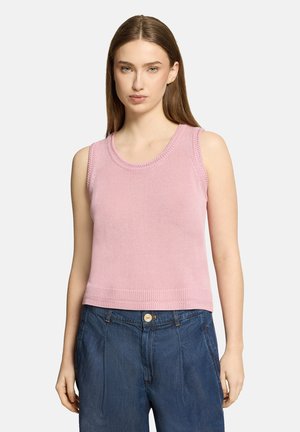 Jeune femme portant un haut en maille rose sans manches et un pantalon en jean bleu taille haute, debout devant un fond clair uni.