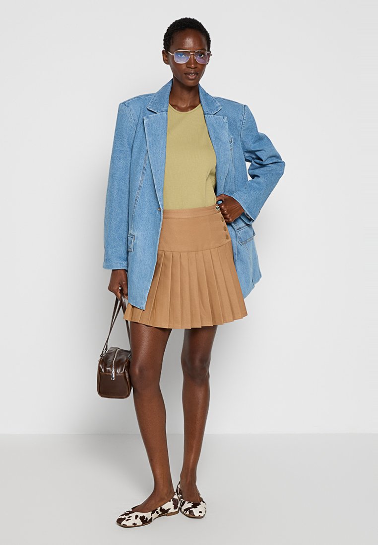 Blazer in denim oversize, top verde, gonna plissettata color sabbia, scarpe a fantasia e borsa marrone. Presenta una vestibilità rilassata e un look a strati.