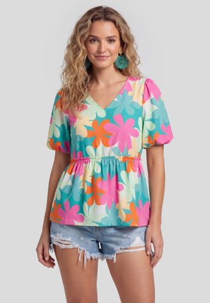 Kobieta w kolorowej, kwiatowej bluzce z krótkimi, bufkowatymi rękawami, w połączeniu z postrzępionymi denimowymi szortami i turkusowymi kolczykami z koralikami.